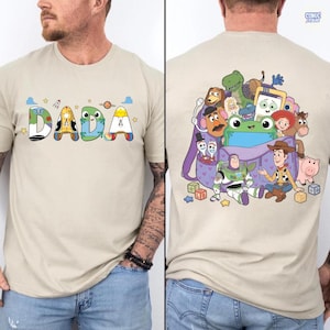 May include: A beige t-shirt featuring the word DADA on the front, with each letter styled after Toy Story characters. The back displays a purple backpack overflowing with various Toy Story characters, including Woody, Buzz Lightyear, and Rex.