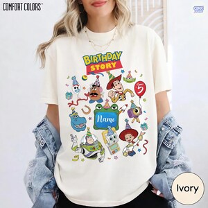 May include: An ivory t-shirt featuring a colorful graphic print with the text "BIRTHDAY STORY" and "Name" in the center. The design includes various cartoon characters wearing party hats, confetti, a red balloon with the number 5, and gifts.