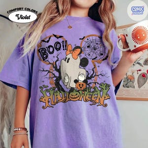 Peut inclure: Un t-shirt violet avec un motif repr&eacute;sentant un fant&ocirc;me portant un n&oelig;ud, tenant une citrouille, entour&eacute; de toiles d'araign&eacute;es, de chauves-souris et d'arbres d&eacute;nud&eacute;s. Le texte indique BOO!! et HALLOWEEN. Le t-shirt est marqu&eacute; COMFORT COLORS Violet.
