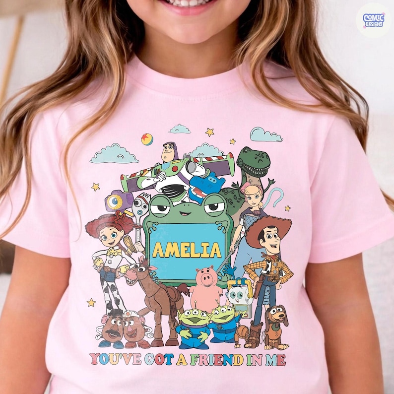 May include: A light pink t-shirt featuring a colorful graphic print of various animated toy characters surrounding a central green frog-shaped frame with the name AMELIA printed inside. The text YOU'VE GOT A FRIEND IN ME is below.