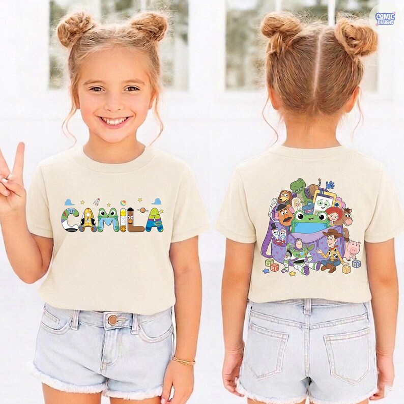 May include: A cream-colored short-sleeve t-shirt featuring the name CAMILA on the front in colorful, character-themed letters. The back displays a graphic of various animated toy characters gathered around a purple backpack with small building blocks.