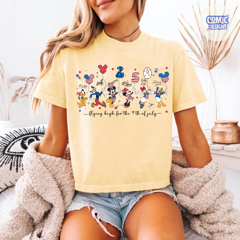 May include: A light yellow t-shirt featuring cartoon characters holding balloons with the numbers 2, 0, 2, and 4. The text "flying high for the 4th of July" is below the characters. The shirt has a crew neck and short sleeves.