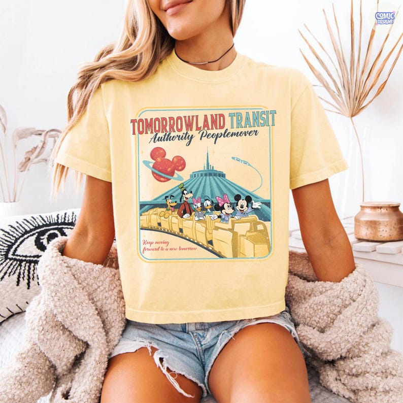 Puede incluir: Camiseta amarilla clara con un dise&ntilde;o gr&aacute;fico retro del paseo Tomorrowland Transit Authority Peoplemover, con Mickey y Minnie Mouse. El dise&ntilde;o incluye el texto "Tomorrowland Transit Authority Peoplemover" y "Keep moving forward to a new tomorrow."