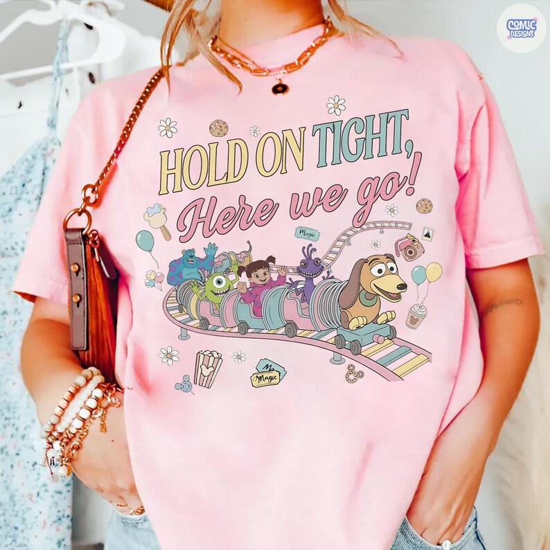May include: Pink t-shirt with the phrase "HOLD ON TIGHT, Here we go!" and a colorful cartoon design featuring characters on a roller coaster. The design includes a dog, monsters, and other whimsical elements. The shirt has a crew neck.