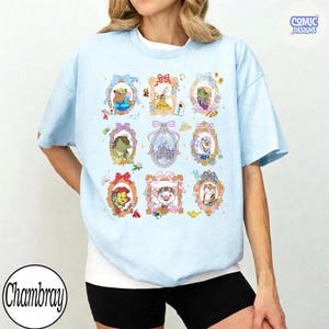 Puede incluir: Camiseta azul claro con retratos enmarcados de personajes de dibujos animados. La camiseta tiene cuello redondo y mangas cortas. El texto "Comic Designs" est&aacute; en la esquina superior derecha y "Chambray" en la esquina inferior izquierda.