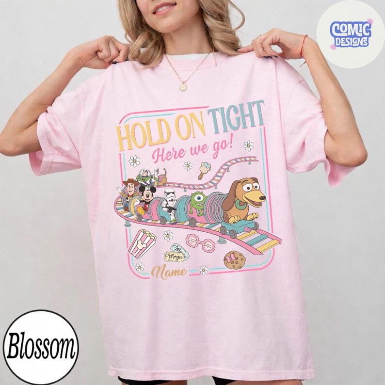 May include: A light pink t-shirt with a colorful graphic featuring cartoon characters on a roller coaster. The text "HOLD ON TIGHT Here we go!" is displayed above the roller coaster design. Additional graphics include popcorn, ice cream, and a cookie.