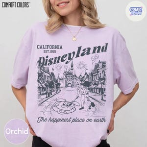 May include: Orchid-coloured t-shirt with a Disneyland graphic. The design features a street scene with a castle, Mickey Mouse, and the text "California Disneyland Est.1955" and "The happiest place on earth". The shirt is from Comfort Colors.