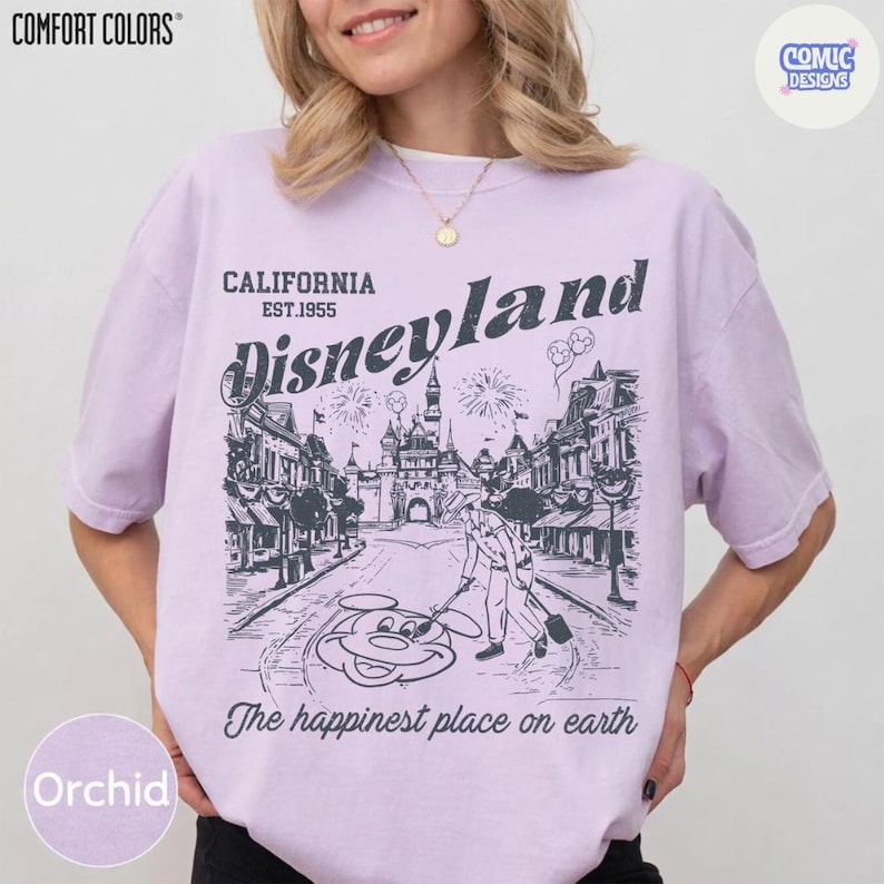 May include: Orchid-coloured t-shirt with a Disneyland graphic. The design features a street scene with a castle, Mickey Mouse, and the text "California Disneyland Est.1955" and "The happiest place on earth". The shirt is from Comfort Colors.