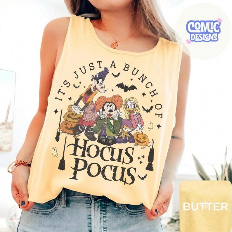 May include: A pale yellow sleeveless tank top featuring a graphic print of three cartoon characters dressed as witches. The text reads "IT'S JUST A BUNCH OF HOCUS POCUS" with illustrations of pumpkins, bats, ghosts, and broomsticks in black ink.