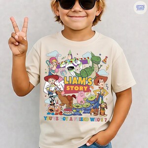 May include: Beige short-sleeve t-shirt featuring a colorful graphic of characters from the animated film "Toy Story." The design includes the text "LIAM'S STORY" and "YOU'VE GOT A FRIEND WHO'S 7?" in a playful font.