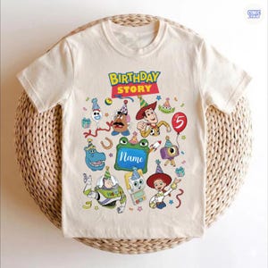May include: A cream-colored short-sleeve t-shirt featuring a colorful graphic print with the text Birthday Story. The design includes various cartoon characters wearing party hats, confetti, a red balloon with the number 5, and a green frame.