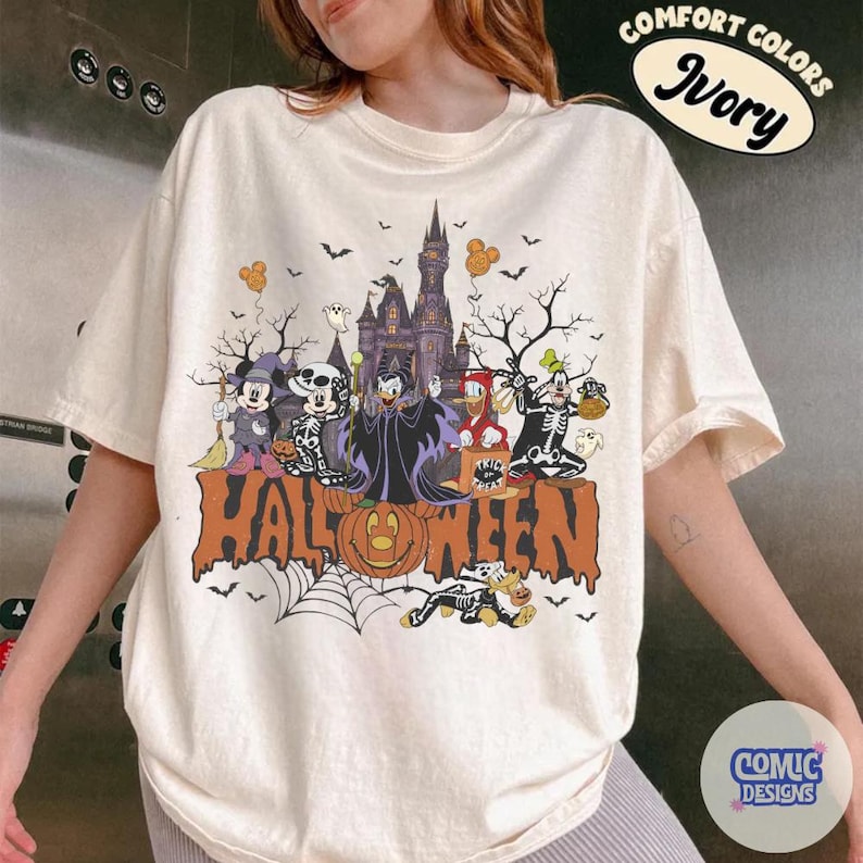 May include: An ivory t-shirt featuring a graphic print of Disney characters in Halloween costumes in front of a castle. The text reads HALLOWEEN, COMFORT COLORS IVORY, and COMIC DESIGNS. The design includes Mickey, Minnie, Goofy, and Donald.