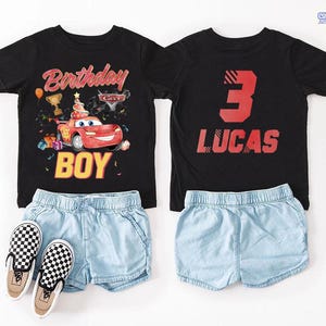 Puede incluir: Camiseta negra con dise&ntilde;o de coche de carreras rojo, con las palabras "Birthday Boy" y "Cars" impresas en la parte delantera. La parte trasera de la camiseta muestra el n&uacute;mero "3" y el nombre "Lucas". Pantalones cortos vaqueros azul claro y zapatos sin cordones a cuadros.