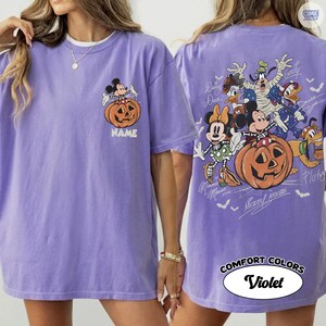 May include: A violet t-shirt featuring a small graphic on the front left chest of Mickey Mouse sitting on a pumpkin with the text NAME. The back displays a large graphic of various cartoon characters in costumes with pumpkins and bats.