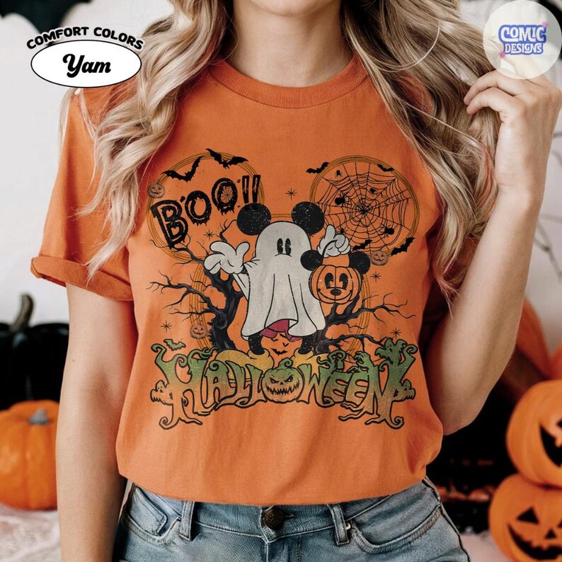 Peut inclure: Un t-shirt orange &agrave; manches courtes avec un motif repr&eacute;sentant un fant&ocirc;me portant des oreilles de souris et tenant un seau citrouille. Le design inclut le texte "BOO!!" et "HALLOWEEN" avec des illustrations de toile d'araign&eacute;e, de chauves-souris et de branches d'arbres d&eacute;nud&eacute;es.