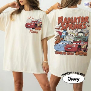May include: An ivory t-shirt featuring a red race car with devil horns and pumpkins on the chest. The back displays multiple cartoon cars in Halloween costumes, spider webs, and the text Radiator Springs Trunk or Treat in orange font.
