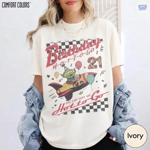 May include: Ivory-colored t-shirt with a retro graphic design. The design features the text "Birthday H-O-T-T-O-GO 21" with a cartoon alien in a rocket ship. The shirt also has the text "You Can Take Me Hot to Go."