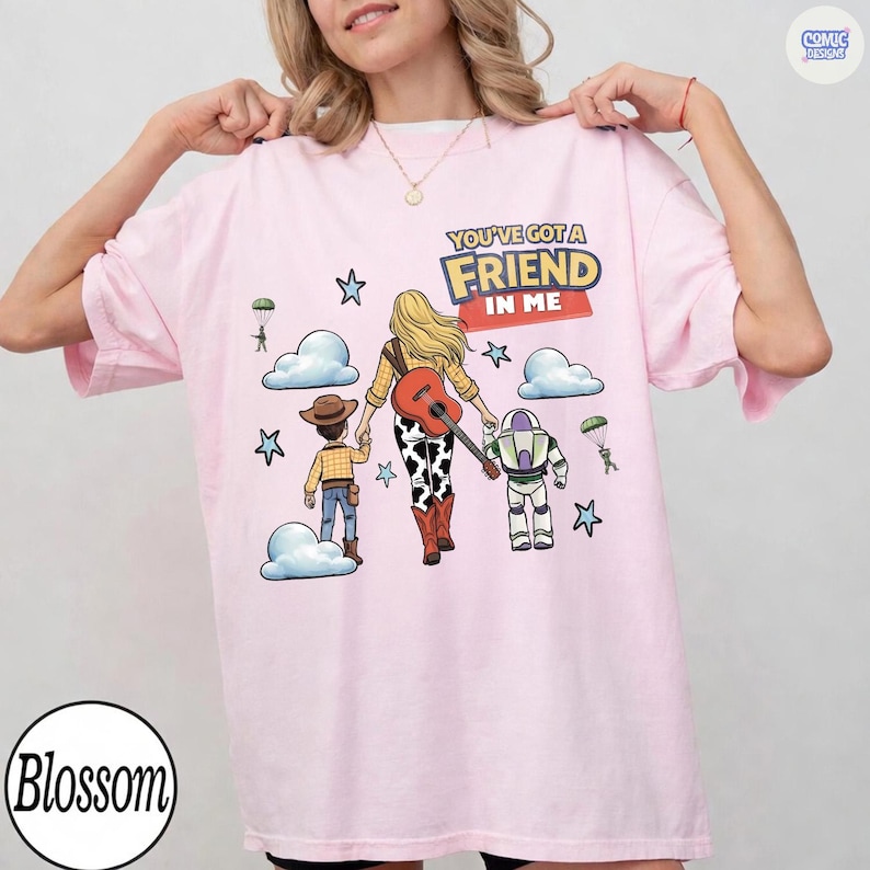 May include: A light pink t-shirt featuring a graphic of a cowboy, a cowgirl with a guitar, and a space ranger character. The text reads YOU'VE GOT A FRIEND IN ME. The design includes blue clouds, stars, and parachuting green soldiers on a light pink background.