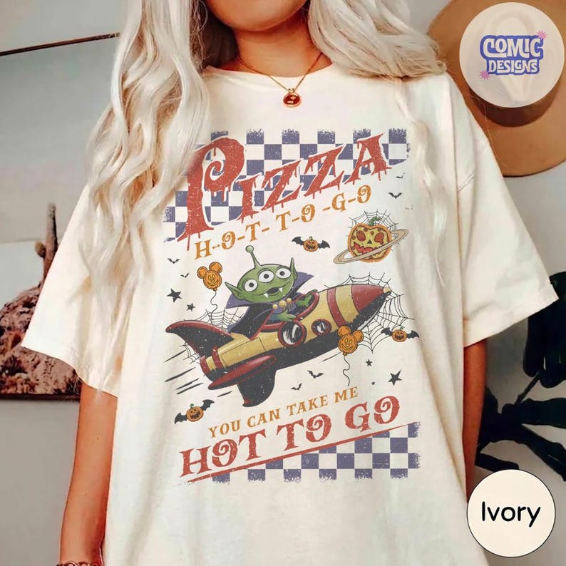 May include: An ivory t-shirt featuring a graphic of a green three-eyed alien in a rocket ship. The design includes a blue and white checkered background, orange text reading Pizza H-O-T-T-O-G-O, and You Can Take Me Hot To Go, with pumpkins.