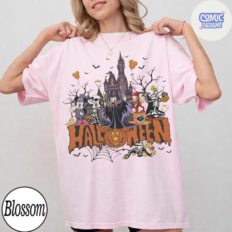 Puede incluir: Una camiseta rosa claro con un estampado gr&aacute;fico de personajes de Disney con disfraces de Halloween frente a un castillo. El texto "HALLOWEEN" est&aacute; impreso en letras grandes de color naranja en la parte inferior del dise&ntilde;o, junto a murci&eacute;lagos, &aacute;rboles y una telara&ntilde;a.