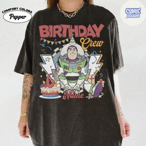 May include: Black Comfort Colors t-shirt featuring a colorful graphic of Buzz Lightyear from Toy Story. The design includes the text "BIRTHDAY Crew" and "6" on a cake, with space for a name. The shirt has a vintage look.