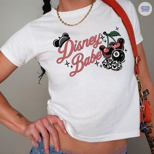 May include: White t-shirt featuring the text "Disney Babe" in pink script, with Mickey Mouse-shaped cherries, an 8-ball, and dice. The design incorporates black accents and star details.