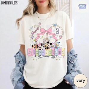 Op de afbeelding: Ivoor kleurig T-shirt met een kleurrijke afbeelding van Disney-figuren, een roze strik en de tekst "Alexcialand". Het ontwerp bevat het woord "Mommy" en het nummer "3". Het shirt is van Comfort Colors en Comic Designs.