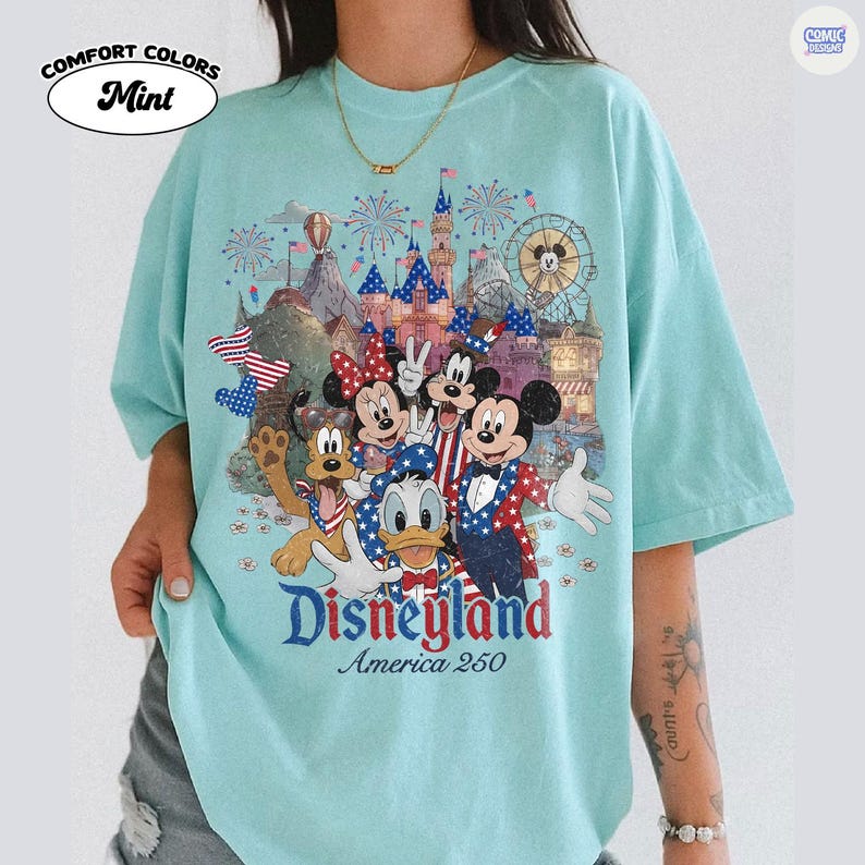 Puede incluir: Una camiseta verde menta con un estampado gr&aacute;fico de Mickey Mouse, Minnie Mouse, Donald Duck, Goofy y Pluto vestidos con ropa patri&oacute;tica roja, blanca y azul. El dise&ntilde;o incluye un castillo, fuegos artificiales y el texto Disneyland America 250.