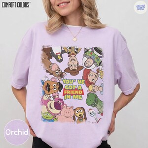 May include: Orchid-colored t-shirt featuring a graphic of characters from the movie Toy Story. The graphic includes Woody, Buzz Lightyear, and other characters with the text "YOU'VE GOT A FRIEND IN ME". The shirt is from Comfort Colors.