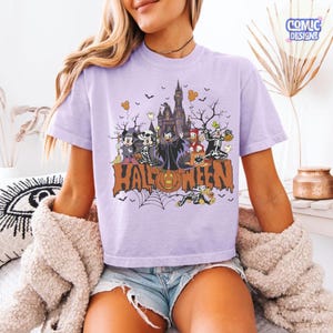 Puede incluir: Una camiseta de color morado claro con un estampado gr&aacute;fico de varios personajes de dibujos animados con disfraces de Halloween, incluyendo un castillo, murci&eacute;lagos y una calabaza. La palabra HALLOWEEN est&aacute; impresa en letras naranjas grandes.