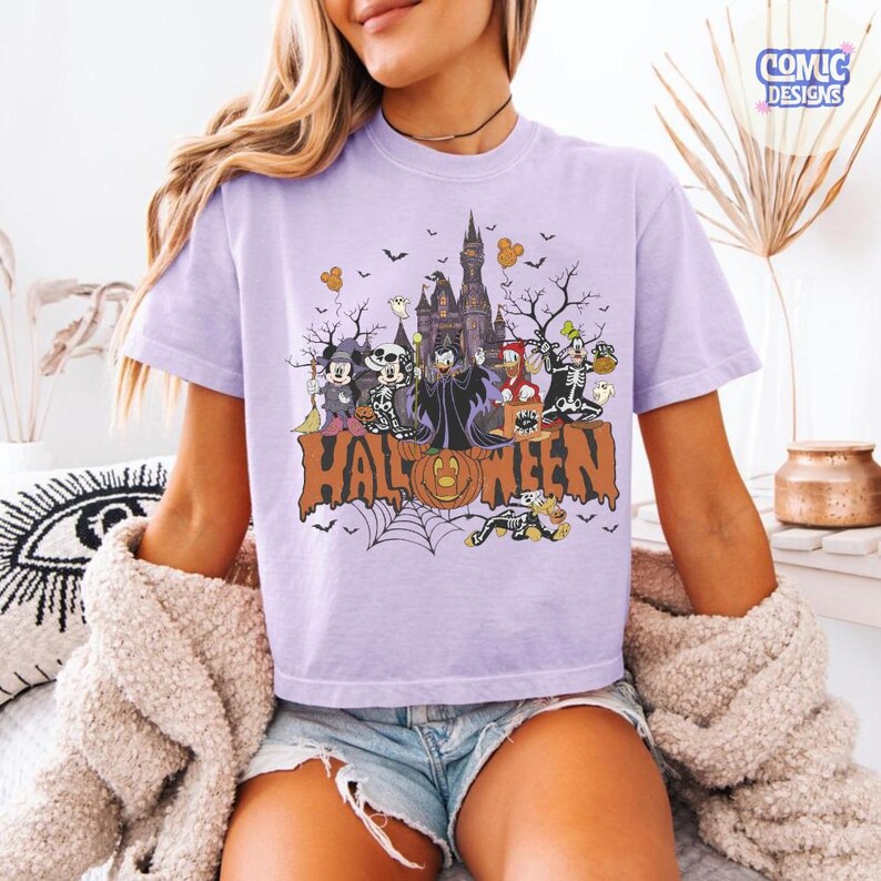 Puede incluir: Una camiseta de color morado claro con un estampado gr&aacute;fico de varios personajes de dibujos animados con disfraces de Halloween, incluyendo un castillo, murci&eacute;lagos y una calabaza. La palabra HALLOWEEN est&aacute; impresa en letras naranjas grandes.