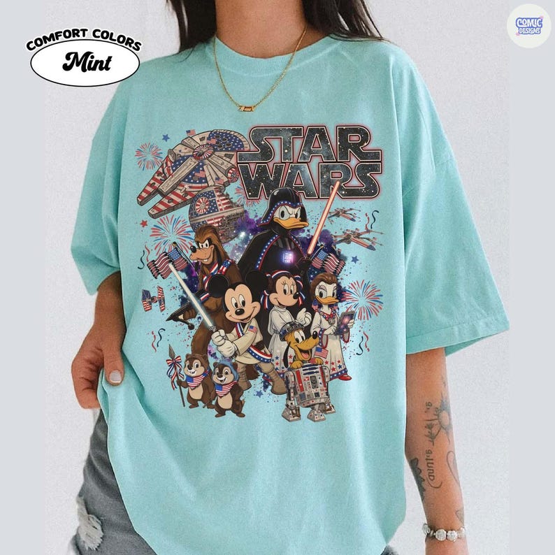 May include: Mint-colored t-shirt featuring a Star Wars graphic with Disney characters and an American flag theme. The design includes the Star Wars logo, a Millennium Falcon, Darth Vader, Mickey Mouse, and Donald Duck. The shirt has the words "Comfort Colors" and "Comic Designs".