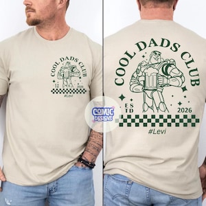 May include: Beige t-shirt with a green graphic of Buzz Lightyear holding a beer mug, with the text "COOL DADS CLUB" and "#Levi". The shirt has a checkered pattern at the bottom and the year "2026".