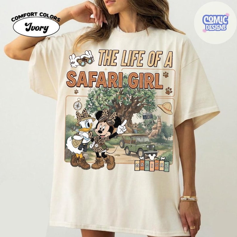 May include: Ivory Comfort Colors t-shirt with a graphic of Minnie and Daisy Duck in safari outfits, with the text "THE LIFE OF A SAFARI GIRL". The design includes a jeep, a tree, and the word "MAGICAL".