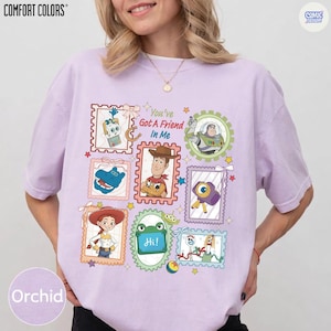 May include: A light purple t-shirt featuring a graphic print of various Toy Story characters arranged in postage stamp-style frames. The text "You've Got A Friend In Me" is printed in the center. The shirt has short sleeves and a crew neckline.