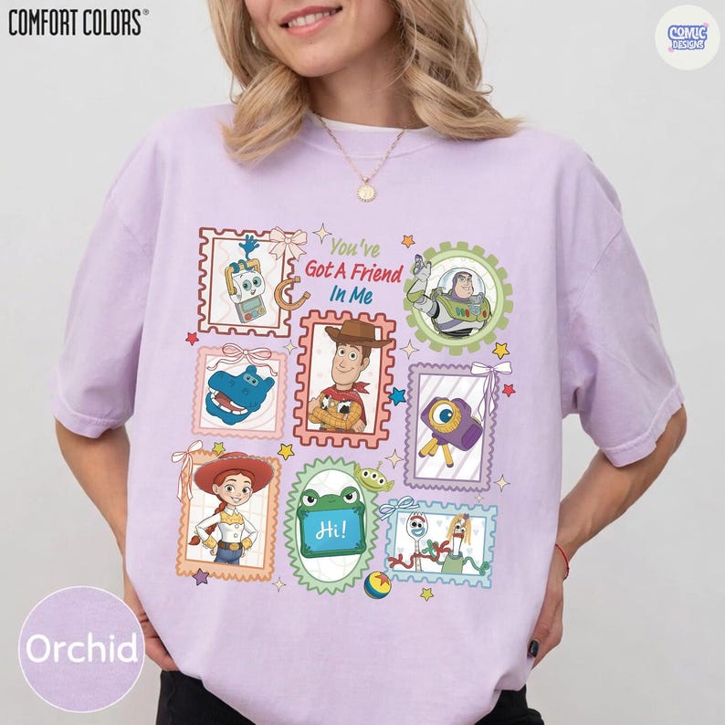 May include: A light purple t-shirt featuring a graphic print of various Toy Story characters arranged in postage stamp-style frames. The text "You've Got A Friend In Me" is printed in the center. The shirt has short sleeves and a crew neckline.