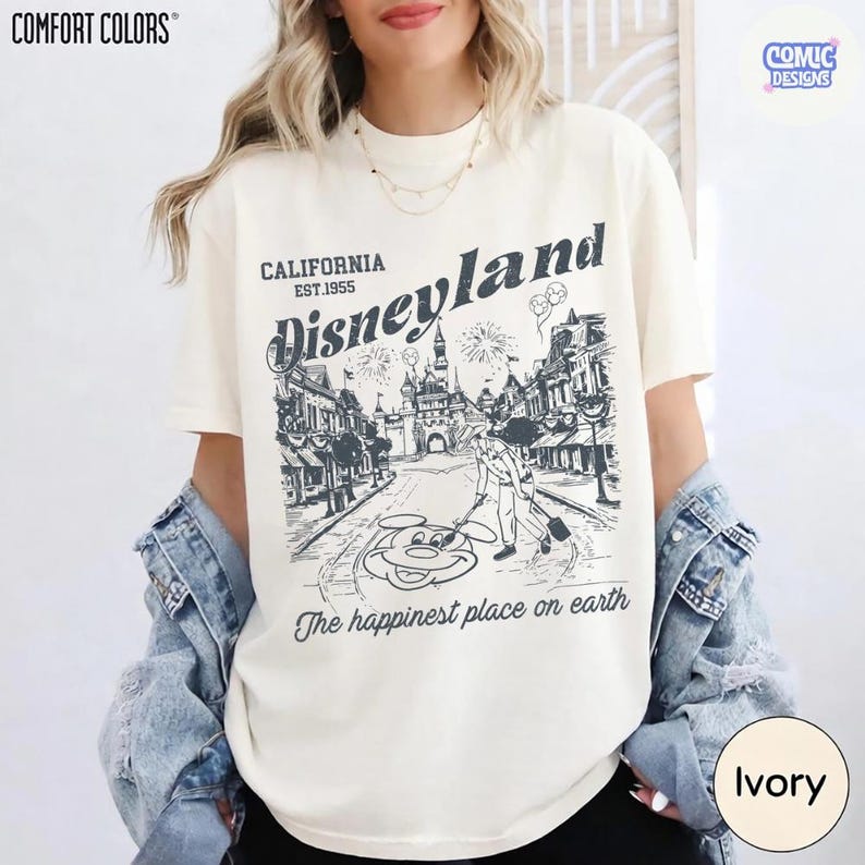 May include: Ivory Comfort Colors t-shirt with a Disneyland graphic. The design features the text "California Est. 1955 Disneyland" and an illustration of the park's entrance and Mickey Mouse. The phrase "The happiest place on earth" is also included.