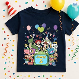 May include: A navy blue short-sleeve t-shirt featuring a colorful graphic of various animated toy characters, balloons, and a banner that reads BIRTHDAY. A central green frog-shaped frame contains the text JAMES and the number 5.