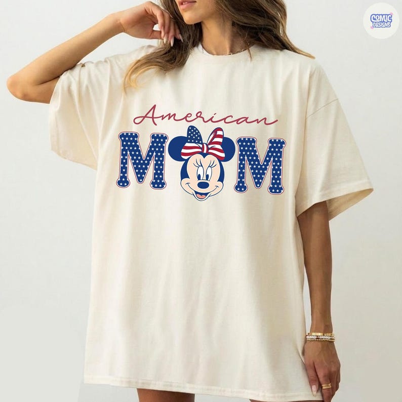 May include: Cream-colored t-shirt featuring the text "American MOM" in red and blue. The letters "MOM" are designed with a star-spangled pattern and include a Minnie Mouse graphic with a patriotic bow.