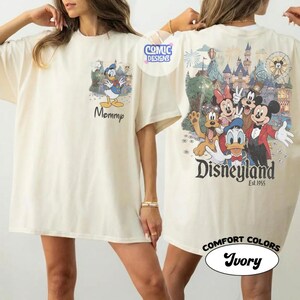 May include: An ivory t-shirt featuring a graphic of Donald Duck on the front chest with the text Mommy. The back displays a large, colorful illustration of Mickey Mouse, Minnie Mouse, Donald Duck, Goofy, and Pluto in front of a castle with Disneyland Est. 1955 text.