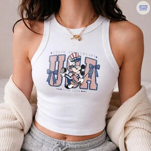 Op de afbeelding: Een wit mouwloos hemdje met een grafische print van Mickey Mouse die een patriottische hoed draagt. De tekst luidt 250TH ANNIVERSARY USA 1776 2026, met rood-wit gestreepte letters en blauwe sterren. Het ontwerp bevat blauwe strikken aan de zijkanten.