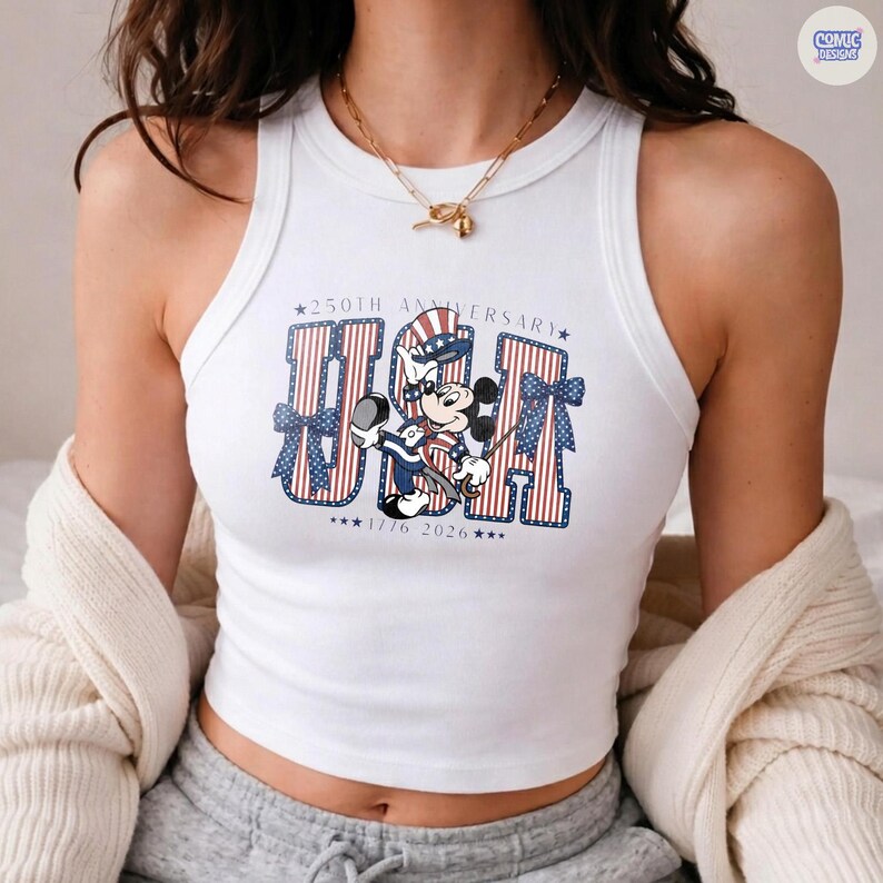 Op de afbeelding: Een wit mouwloos hemdje met een grafische print van Mickey Mouse die een patriottische hoed draagt. De tekst luidt 250TH ANNIVERSARY USA 1776 2026, met rood-wit gestreepte letters en blauwe sterren. Het ontwerp bevat blauwe strikken aan de zijkanten.