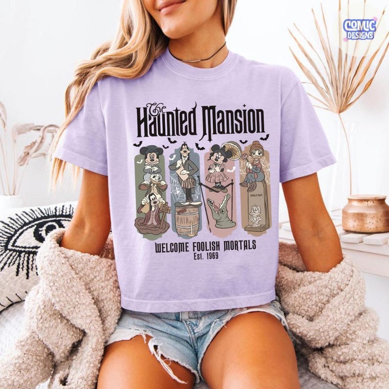 May include: A light purple t-shirt featuring the text "Haunted Mansion" in a gothic font, with cartoon characters and the phrase "Welcome Foolish Mortals Est. 1969." The design includes bats and various themed illustrations.