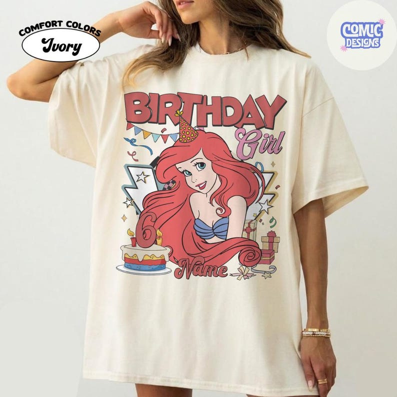 May include: Ivory-colored t-shirt with a graphic of a cartoon mermaid with red hair, wearing a blue top and a party hat. The text "BIRTHDAY Girl" is above the mermaid, and a cake with a number 6 is below.