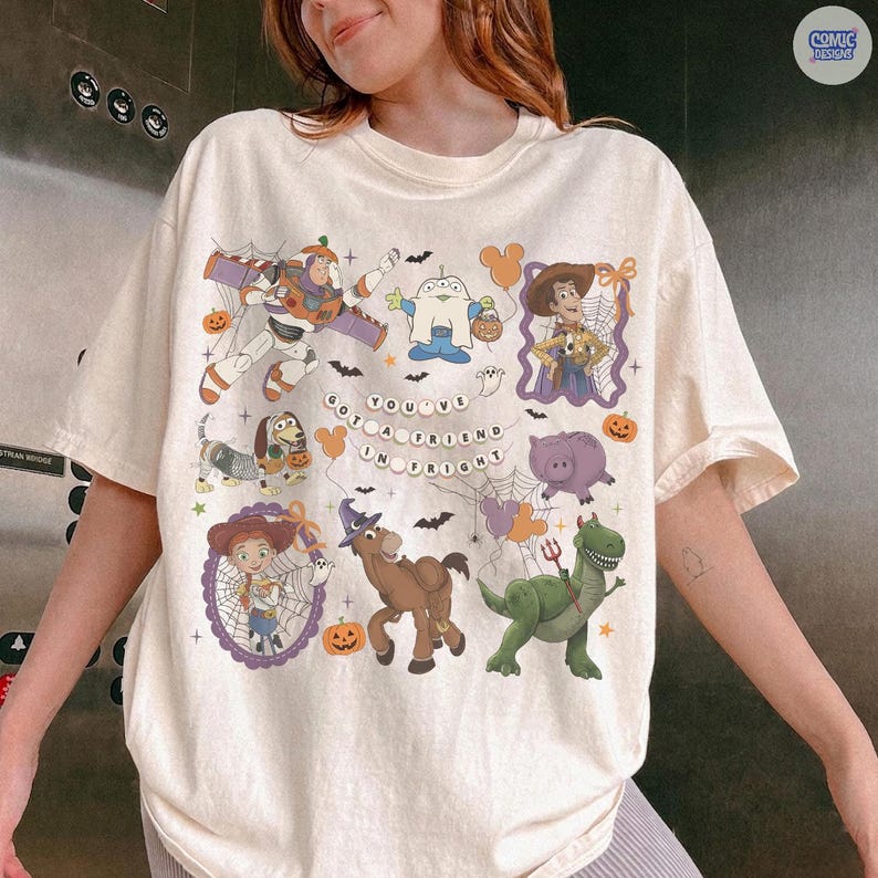 May include: A cream-colored t-shirt featuring a Halloween-themed graphic with Toy Story characters including Buzz Lightyear, Woody, Jessie, Rex, and Slinky Dog. The design includes spiderwebs, pumpkins, and the text 'YOU'VE GOT A FRIEND IN FRIGHT'.