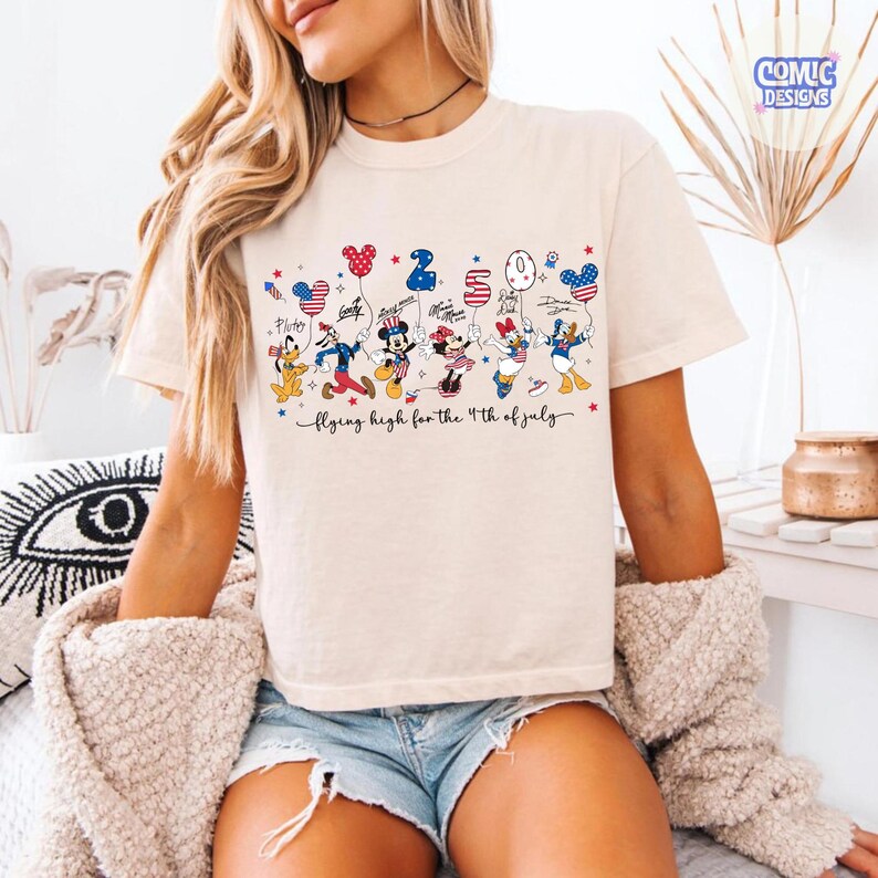 May include: Cream-colored t-shirt featuring a graphic of Disney characters holding balloons with patriotic designs. The text "Flying high for the 4th of July" is written in cursive below the characters. The shirt has short sleeves and a crew neck.