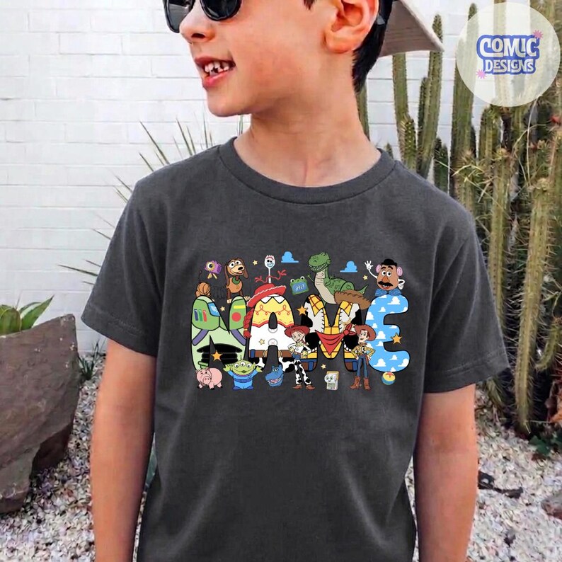 May include: A dark grey short-sleeve t-shirt featuring a colourful graphic print of various animated characters arranged to spell out the word NAME. The design includes figures like a space ranger, a cowboy, a dinosaur, and a potato-shaped character.