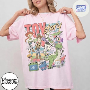 May include: A light pink short-sleeve t-shirt featuring a graphic print of Toy Story characters including Jessie, Woody, Buzz Lightyear, Rex, and others. The design includes the text Toy Story, Hi, and various colorful shapes and star accents.