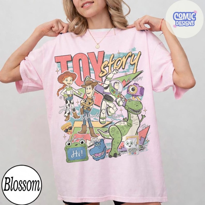 May include: A light pink short-sleeve t-shirt featuring a graphic print of Toy Story characters including Jessie, Woody, Buzz Lightyear, Rex, and others. The design includes the text Toy Story, Hi, and various colorful shapes and star accents.