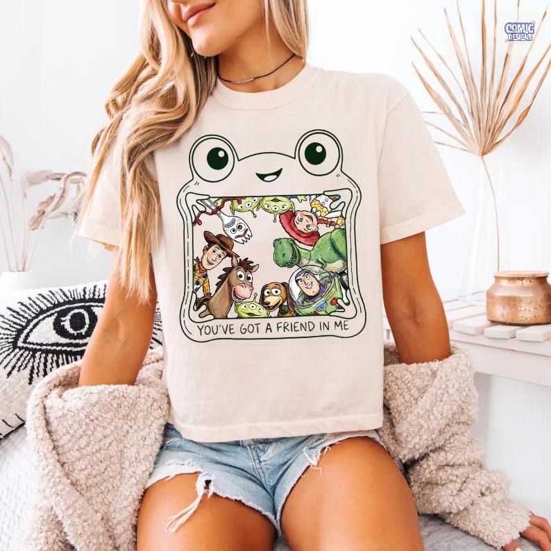 May include: A cream-colored t-shirt featuring a graphic of a frog-shaped frame containing various animated characters from the Toy Story franchise. Below the characters, the text reads, YOU'VE GOT A FRIEND IN ME, in a simple black sans-serif font.